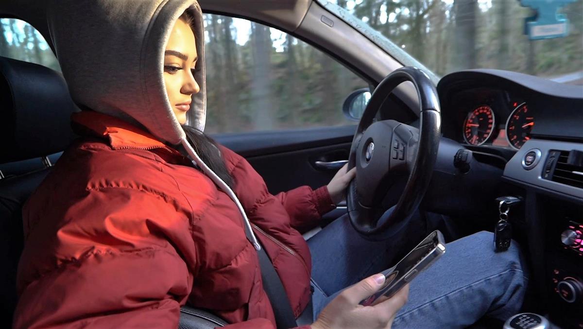 Ung kvinne bak rattet i bil med mobil i hånden. - Klikk for stort bilde